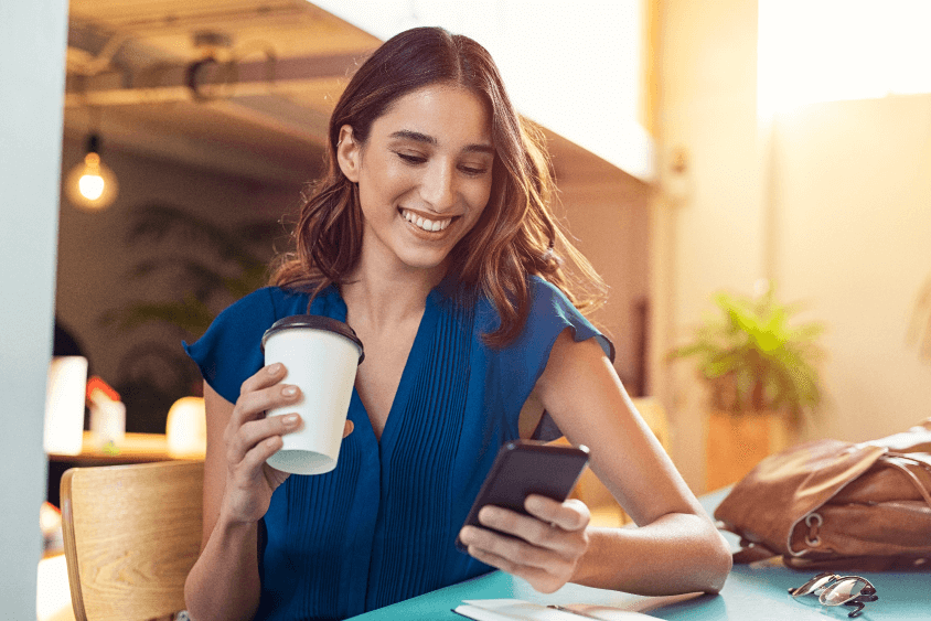 Young, brunette woman smiling holding a coffee and looking at her phone